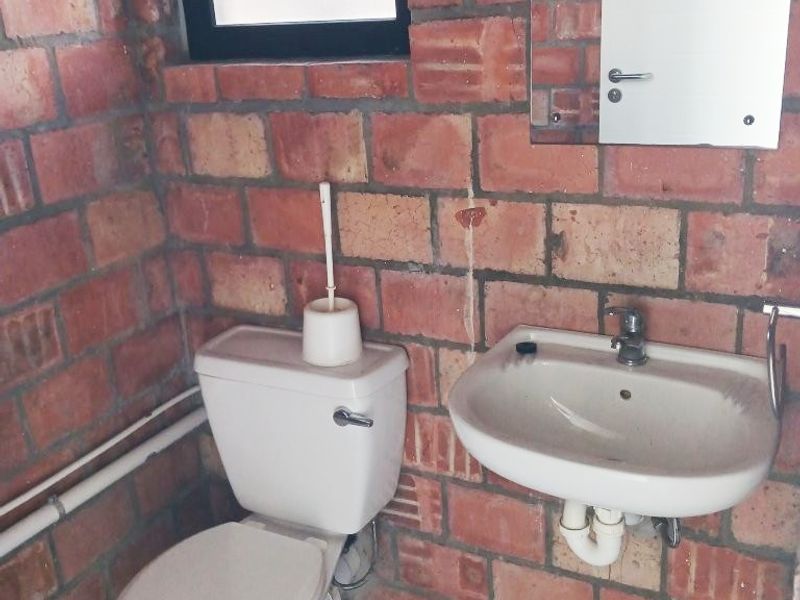 A compact office bathroom featuring exposed brick walls, a wall-mounted basin, and a standard toilet, requiring cosmetic updates to improve the finish.