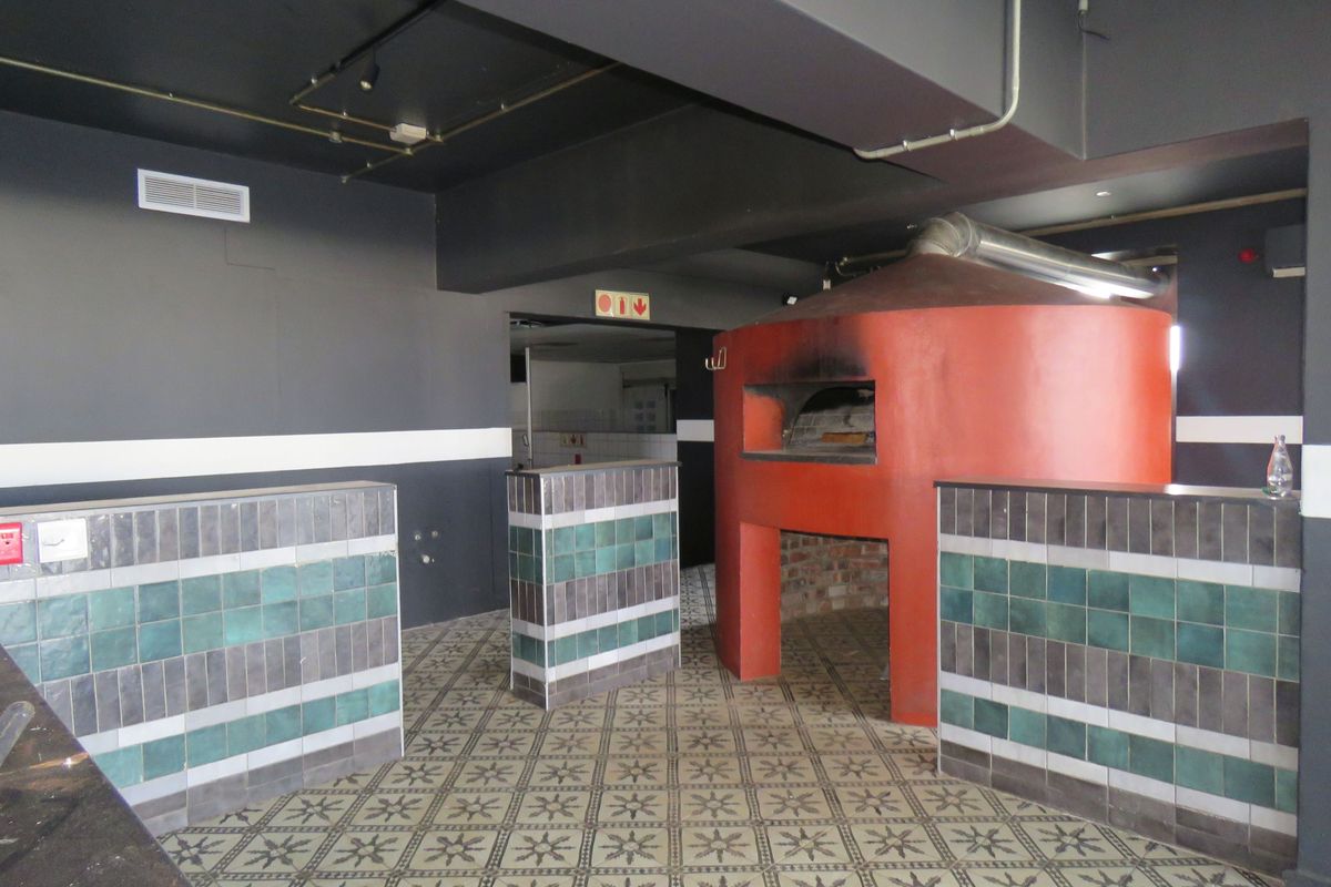 Commercial kitchen area featuring a prominent built-in pizza oven, decorative tiled counters, and patterned floor tiles in a modern industrial setting.