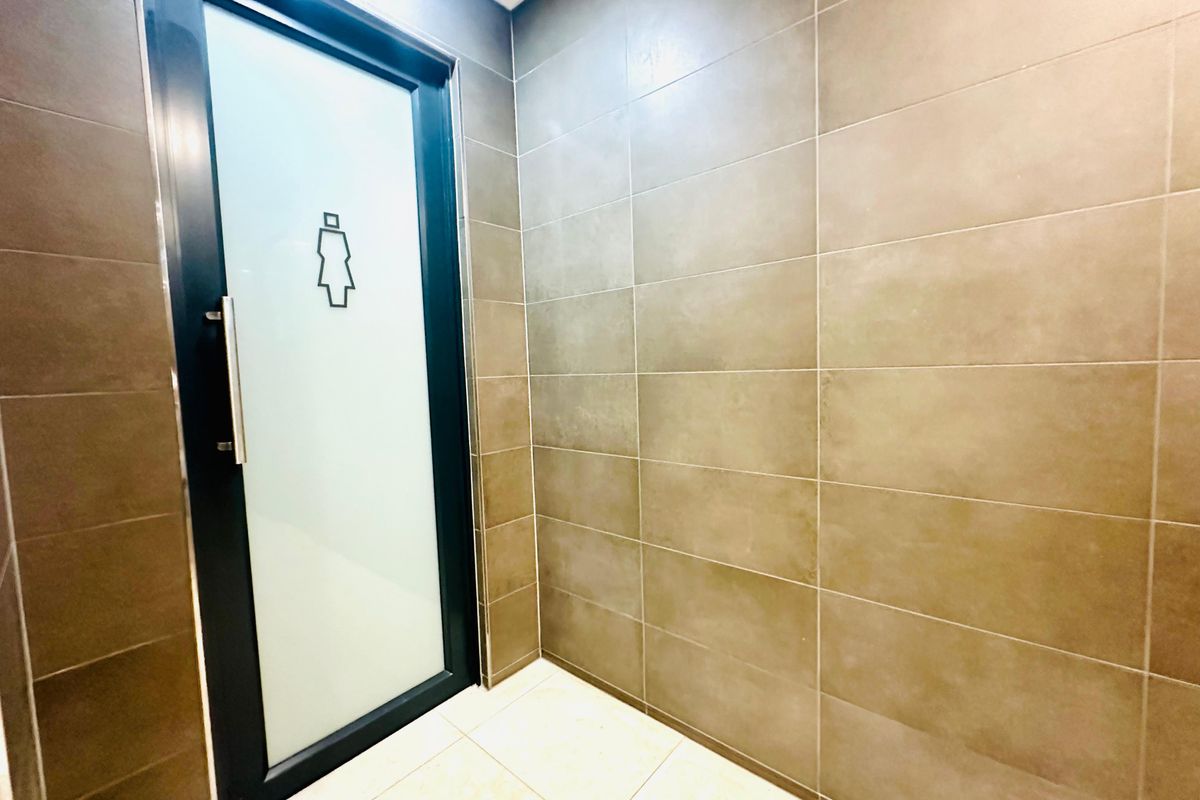 A modern office restroom entrance featuring large-format brown wall tiles, light-colored floor tiles, and a frosted glass door with signage.