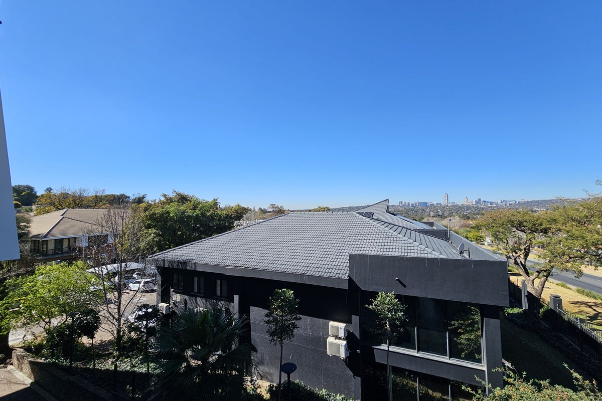 Elevated aerial view of a modern office building with a dark exterior finish, tiled roof, and surrounding landscaped grounds with city skyline views.