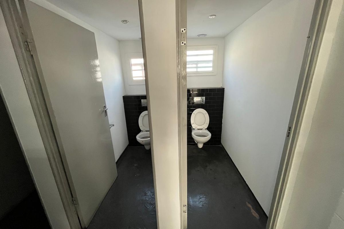 Two individual restroom cubicles featuring wall-mounted toilets, dark tiled splashbacks, and louvered windows within an industrial facility.