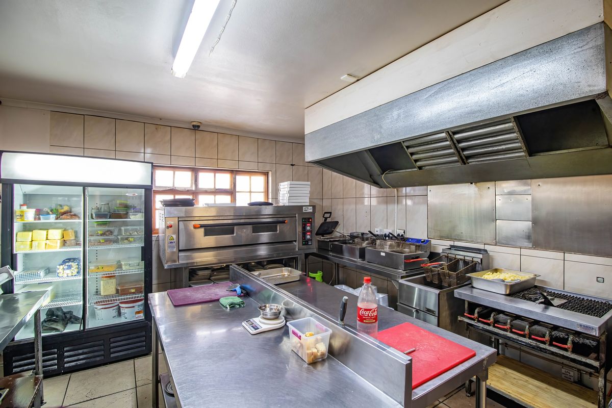Commercial kitchen featuring stainless steel prep tables, industrial extractor hood, commercial oven, and deep fryers with tiled wall finishes.