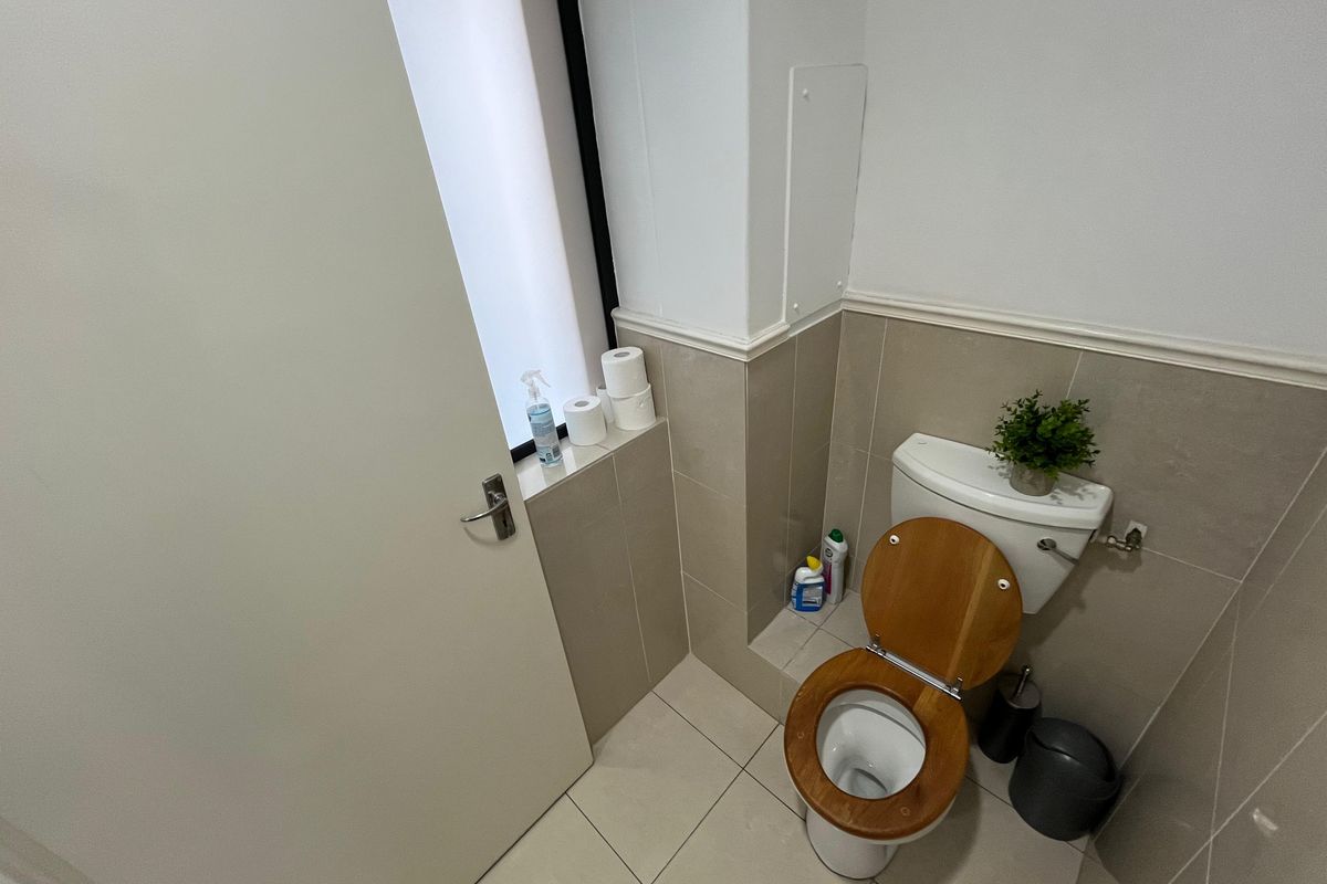 A compact office restroom featuring neutral wall tiling, tiled flooring, and a modern toilet suite with a wooden seat.