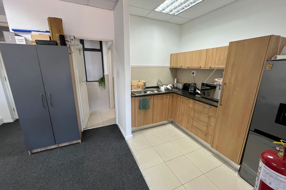 A compact office kitchenette featuring light wood cabinetry, a stainless steel sink, and tiled flooring, adjacent to a carpeted office workspace.