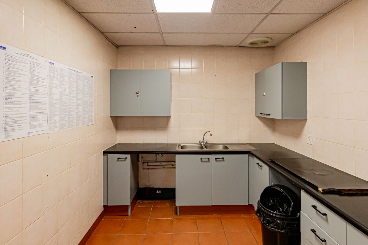 A functional office kitchenette featuring tiled walls, built-in cabinetry, a stainless steel sink, and a drop ceiling with integrated lighting.