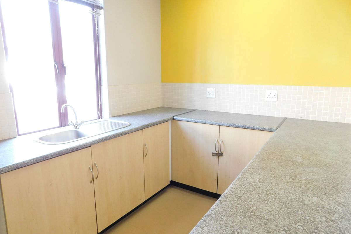 A functional office kitchenette featuring light wood cabinetry, granite-look countertops, a stainless steel sink, and tiled splashbacks against a bright yellow accent wall.