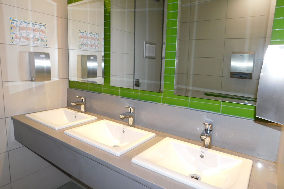 A modern office restroom vanity area featuring three integrated white ceramic basins, chrome faucets, and vibrant green tiled backsplash with large wall-mounted mirrors.
