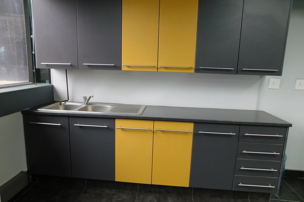 A modern office kitchenette featuring two-tone grey and yellow cabinetry, a stainless steel sink, and dark tiled flooring.