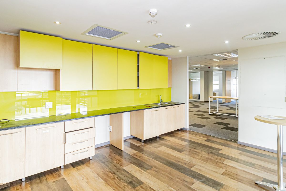 A modern office kitchenette featuring light wood cabinetry, a vibrant yellow glass splashback, dark stone countertops, and recessed ceiling lighting.