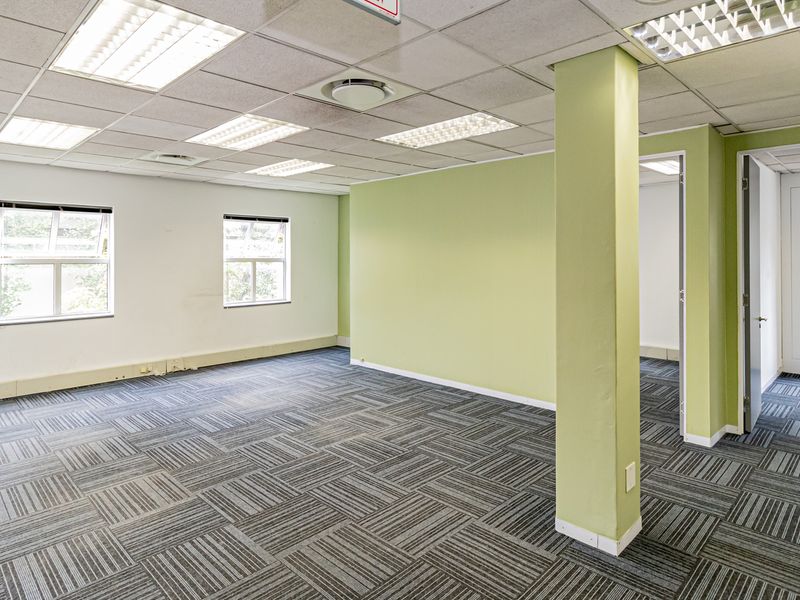 A spacious, empty office area featuring carpet tiles, a suspended ceiling with recessed lighting, and a central structural column.