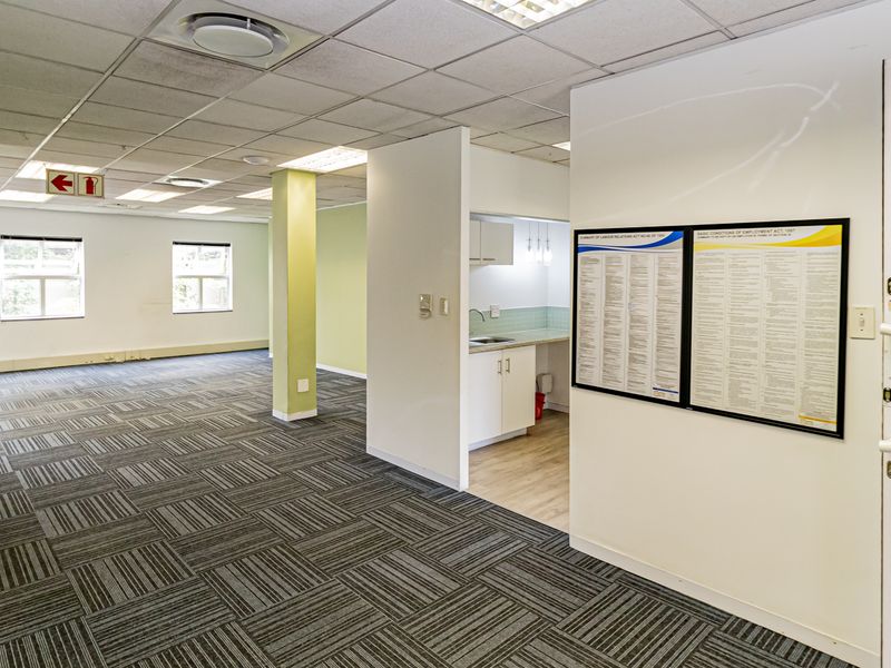 Open-plan office space featuring carpet tiles, a suspended ceiling, and a small integrated kitchenette area with built-in cabinetry and sink.
