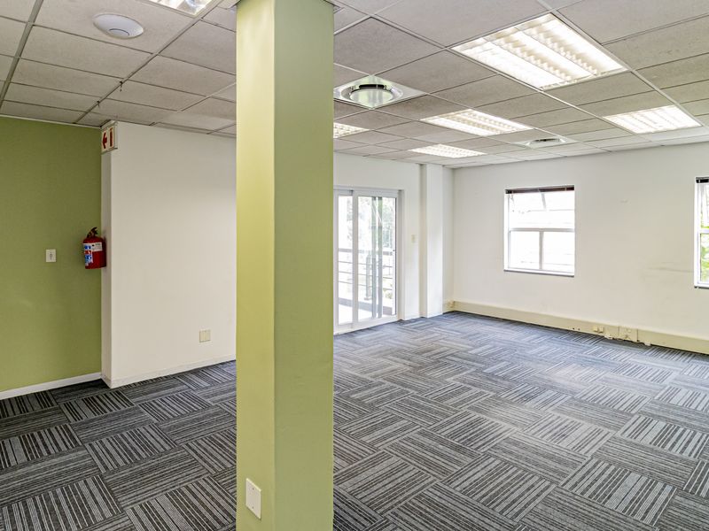 A spacious, empty office area featuring carpet tiles, a suspended ceiling with recessed lighting, and a sliding glass door leading to an exterior balcony.