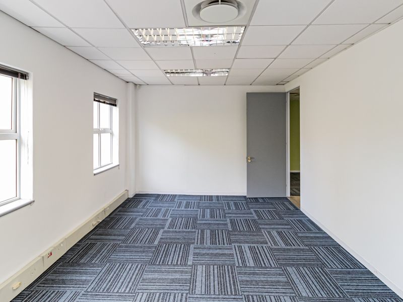 A clean, empty office space featuring carpet tiles, suspended ceilings with recessed lighting, and neutral white walls, ready for professional use.