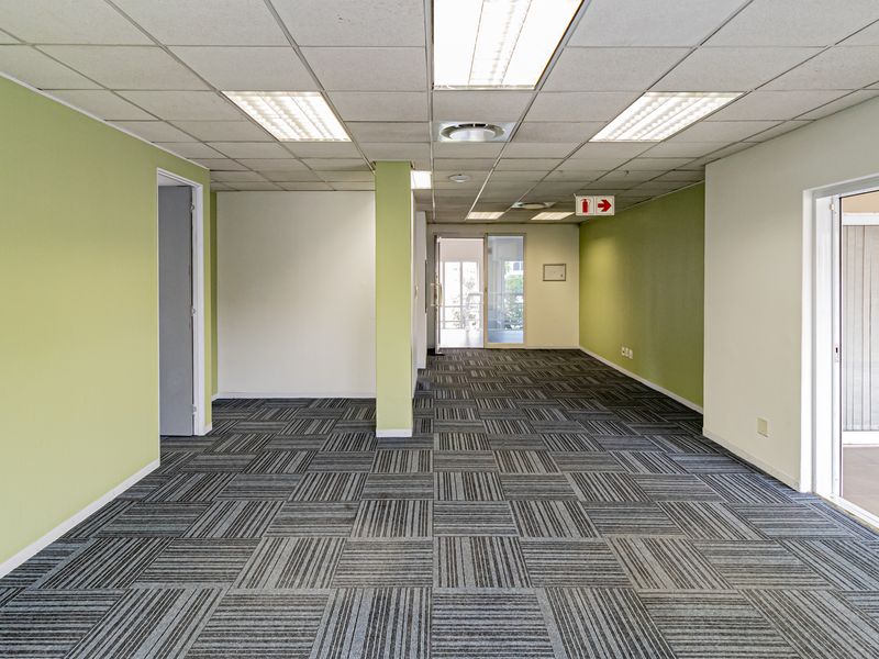 A spacious, empty office area featuring carpet tiles, a suspended ceiling with recessed lighting, and a sliding glass door leading to an outdoor space.