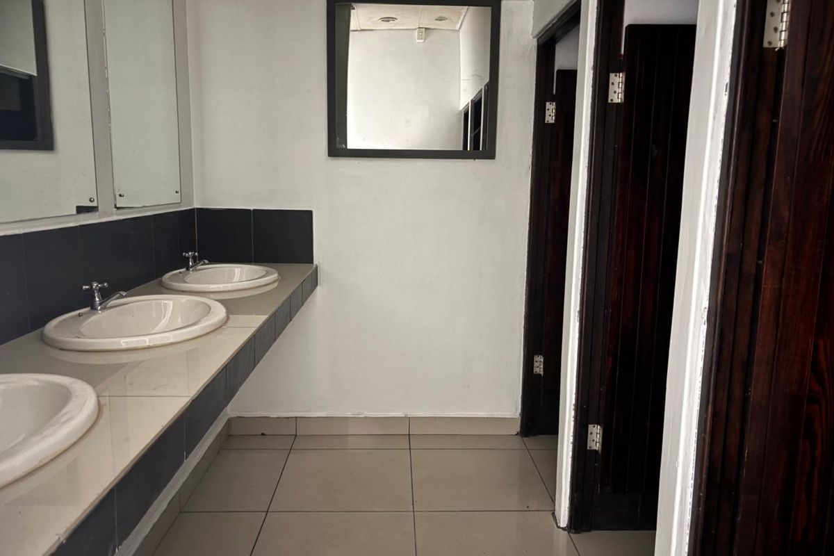 A functional commercial bathroom featuring a long vanity with three integrated washbasins, tiled splashbacks, and multiple private stall entrances.