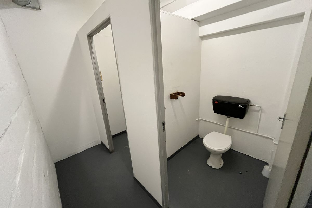 A functional industrial-style restroom featuring a basic toilet suite, partition walls, and exposed plumbing, suitable for warehouse or workshop use.