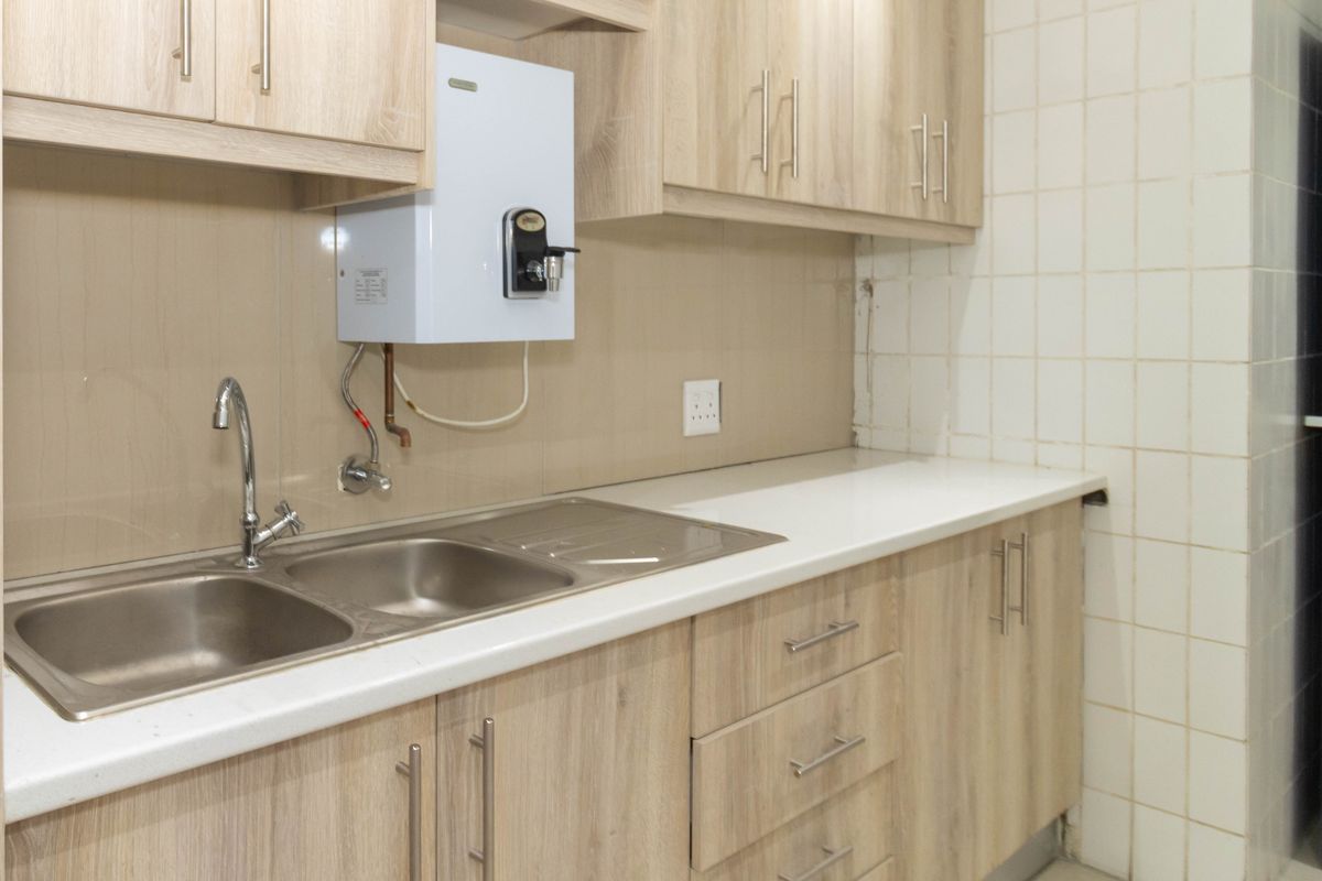 A functional kitchenette area featuring light wood-grain cabinetry, a stainless steel double sink, and a wall-mounted water boiler unit.