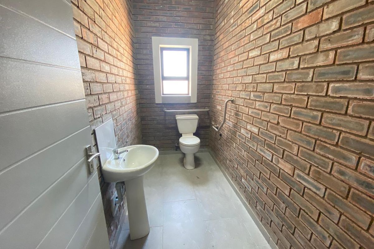 A functional industrial-style bathroom featuring exposed brick walls, a pedestal basin, a toilet, and stainless steel grab rails for accessibility.