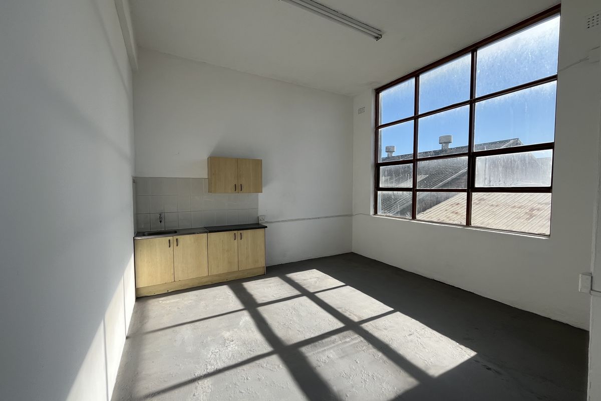 A functional industrial kitchenette featuring light wood cabinetry, a stainless steel sink, and large industrial-style windows providing ample natural light.