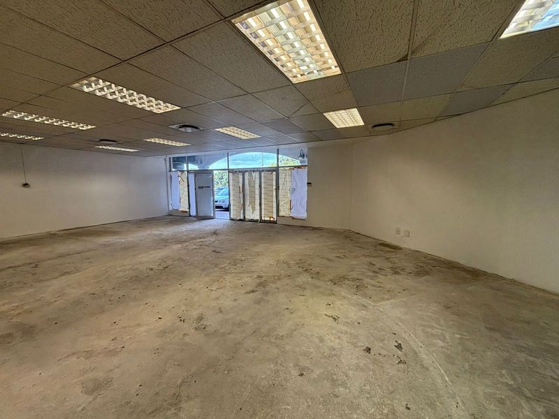 A vacant open-plan retail space featuring a suspended ceiling with recessed lighting, large glass storefront windows, and unfinished concrete flooring.