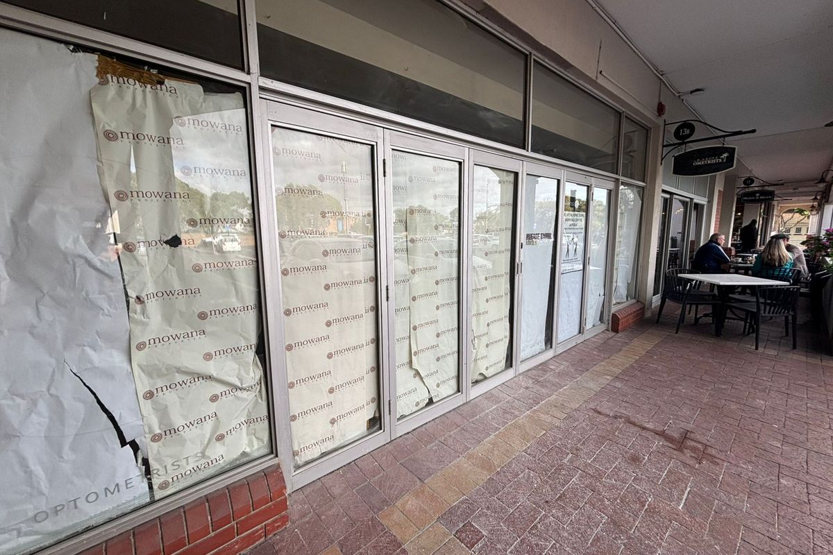 Retail storefront featuring large glass windows with branded film, situated along a paved covered walkway in a commercial strip.