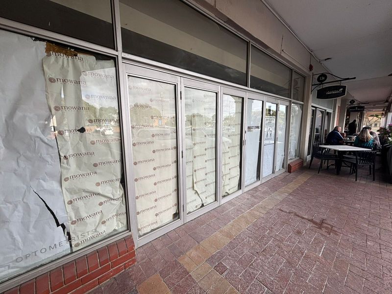 Retail storefront featuring large glass windows with branded film, situated along a paved covered walkway in a commercial strip.