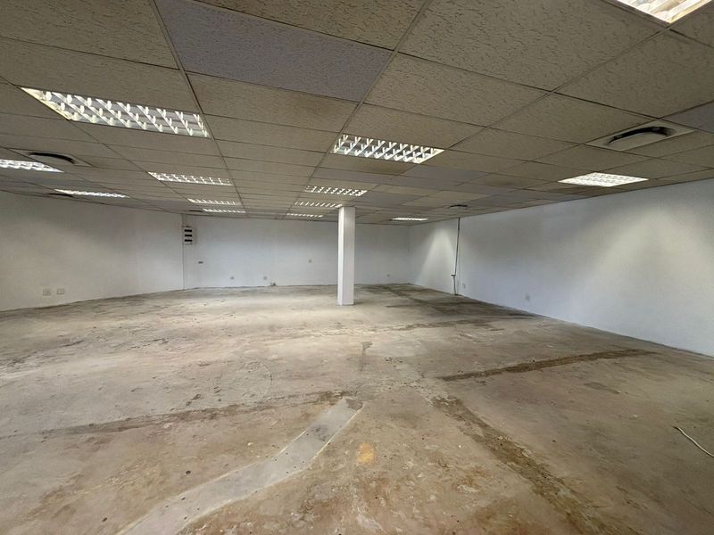 An expansive, empty open-plan retail space featuring a suspended ceiling with recessed lighting, a central support column, and unfinished concrete flooring.