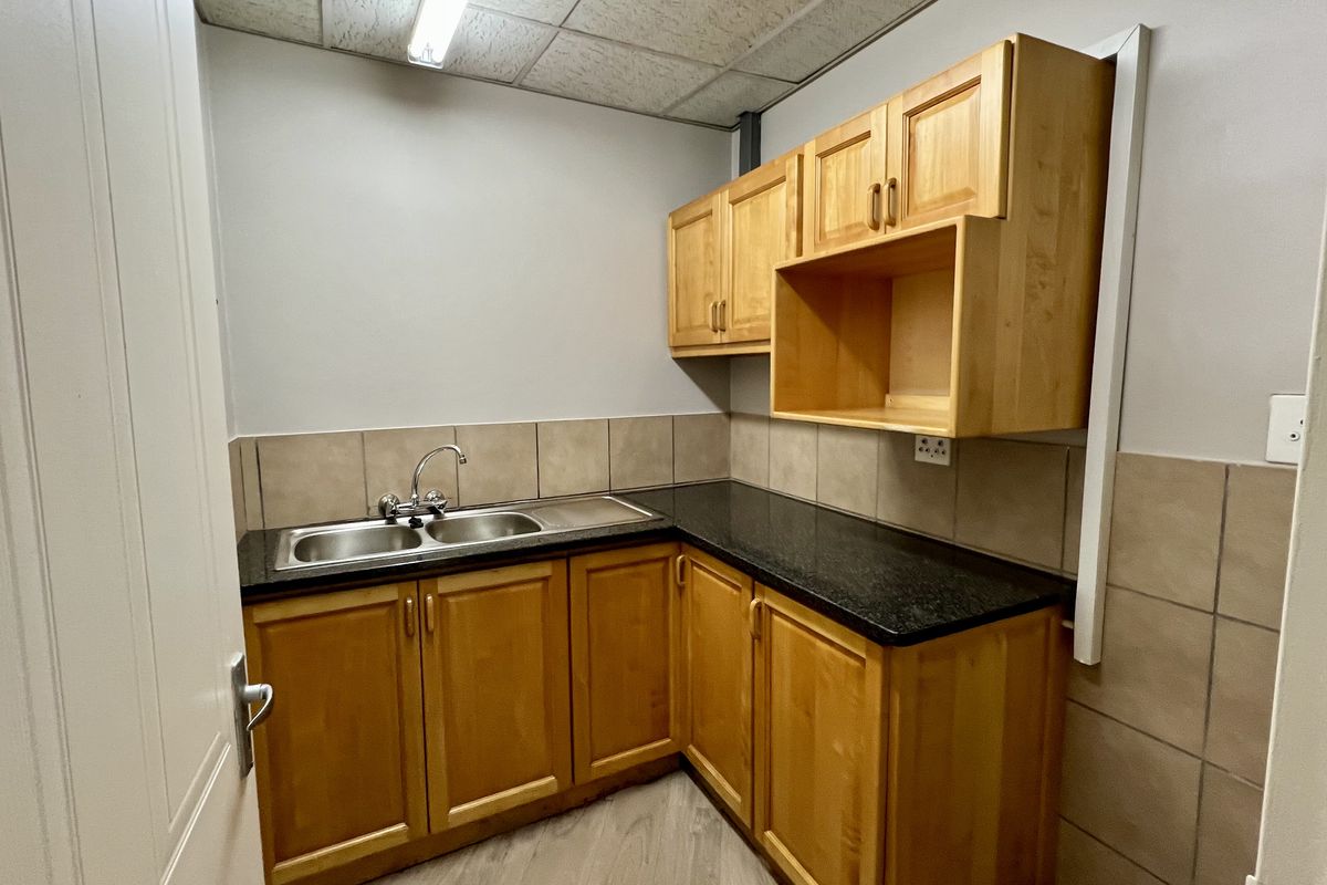 A functional office kitchenette featuring wooden cabinetry, a dark stone-look countertop, a double stainless steel sink, and tiled splashbacks.