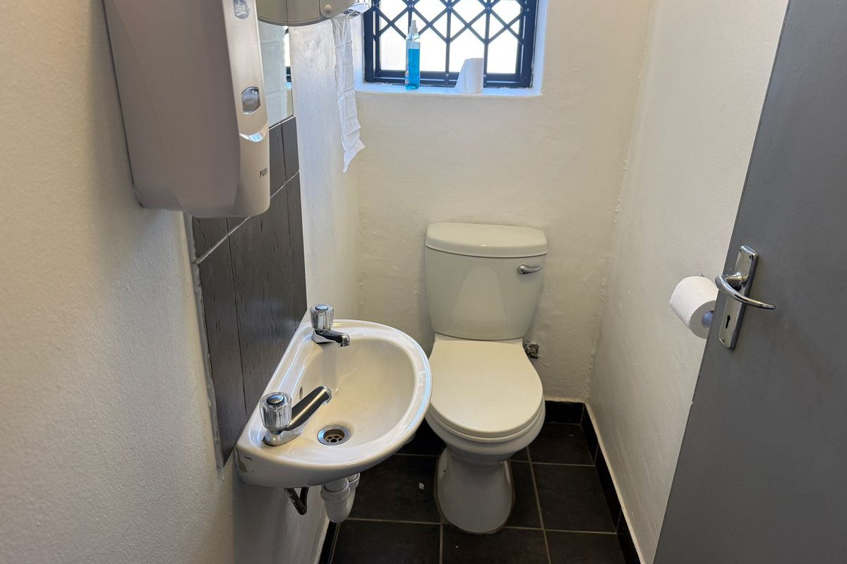 Compact industrial restroom featuring a wall-mounted basin, standard toilet, tiled flooring, and a security-grille window.