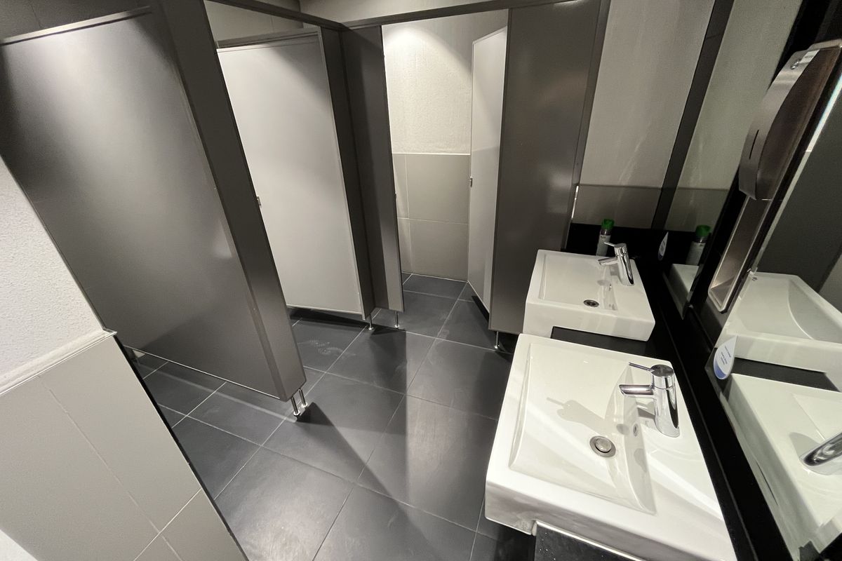 Modern office restroom featuring dual wall-mounted ceramic basins, dark tiled flooring, and partitioned toilet cubicles with a clean, professional finish.