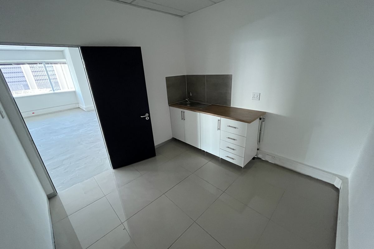 A compact office kitchenette featuring white cabinetry, a wooden countertop, a stainless steel sink, and tiled flooring, adjacent to an open office space.