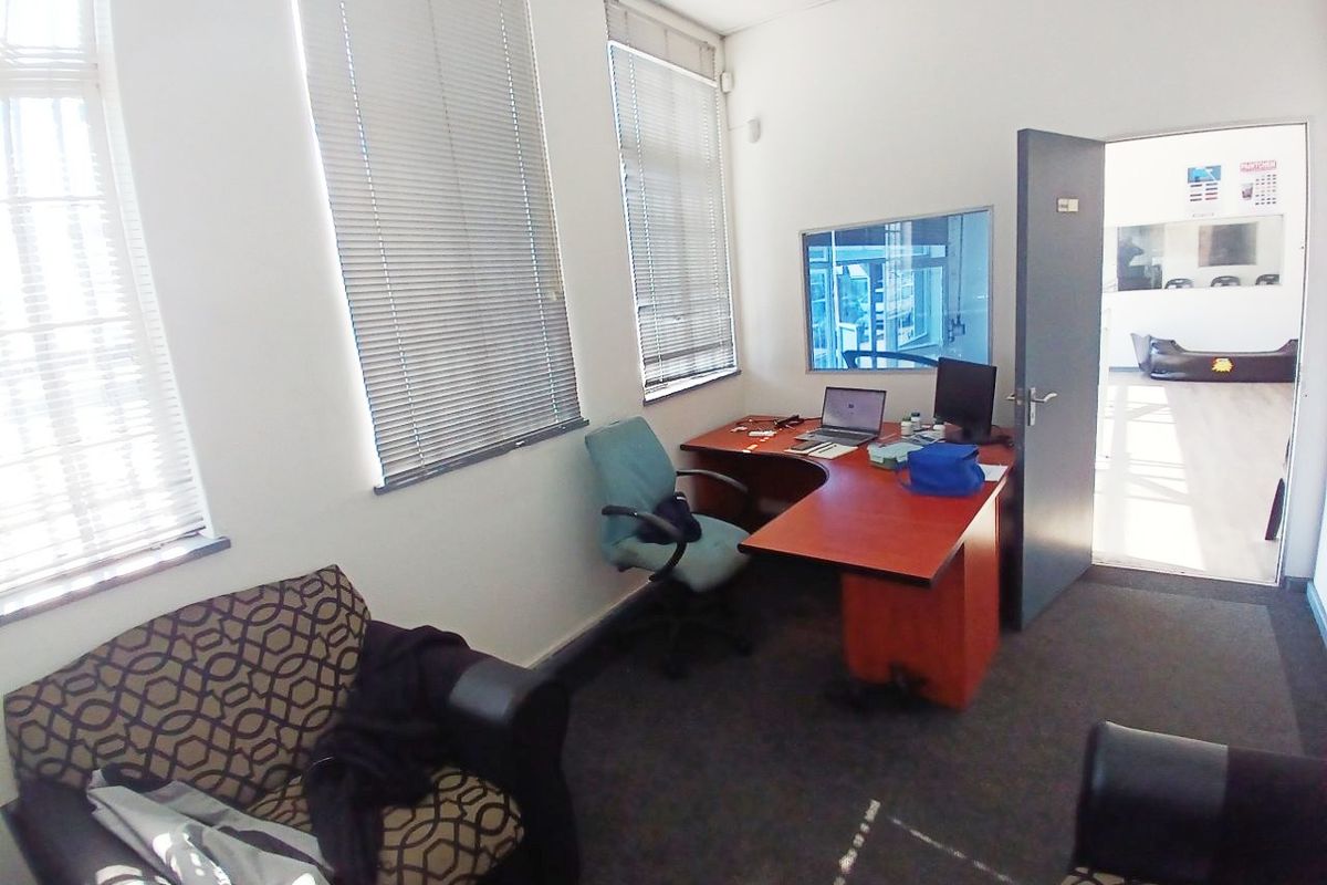 A functional office space featuring large windows with fitted blinds, carpeted flooring, and an internal viewing window looking into the adjacent area.