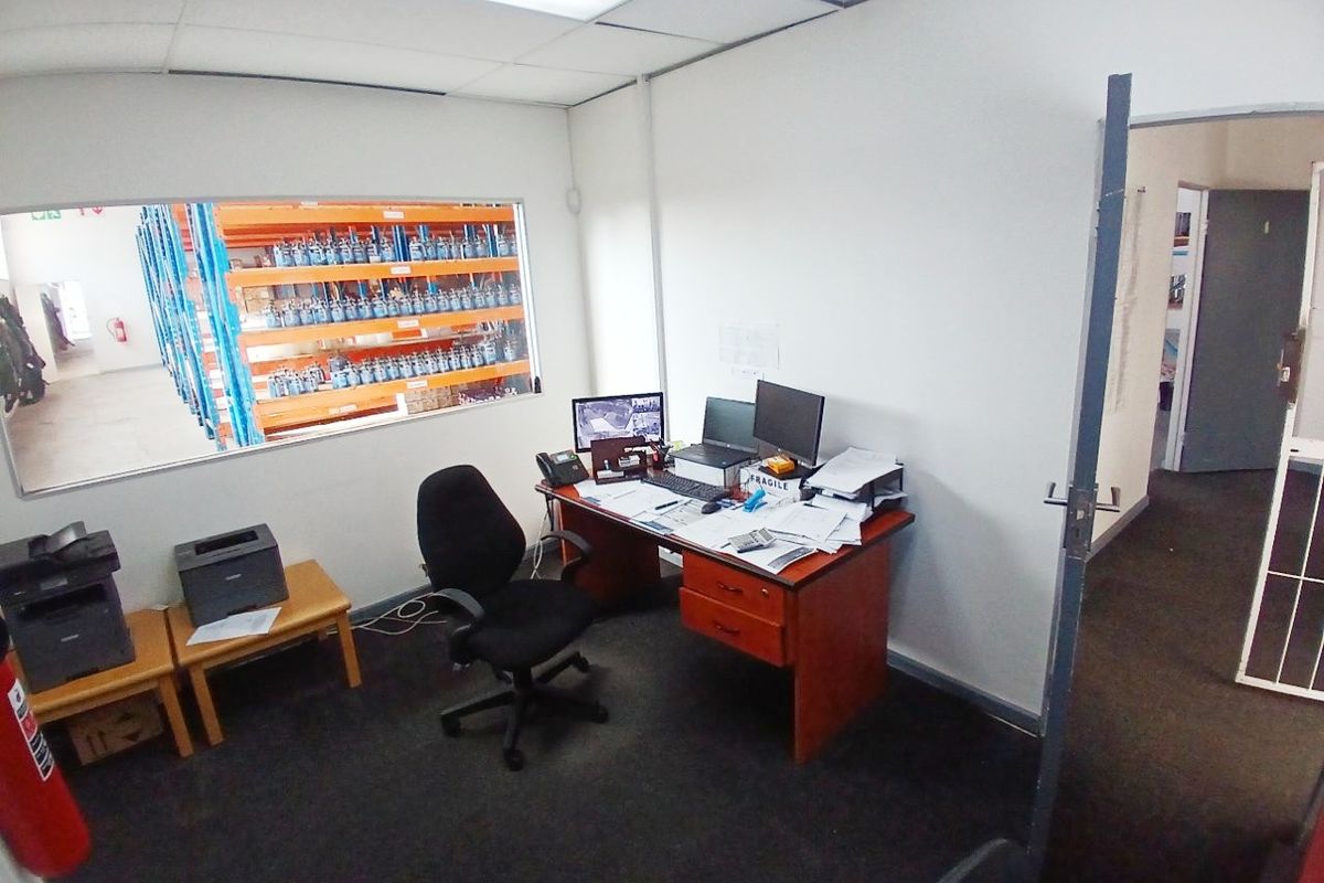 A functional industrial office space featuring a large viewing window overlooking the warehouse floor, suspended ceiling tiles, and carpeted flooring.