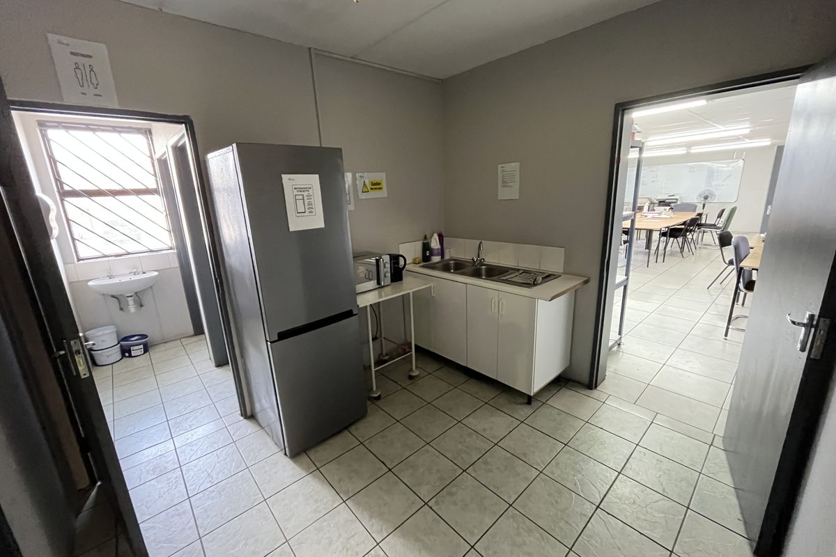 A functional industrial kitchenette area featuring a stainless steel sink, base cabinetry, and tiled flooring, with an adjacent restroom and office space access.