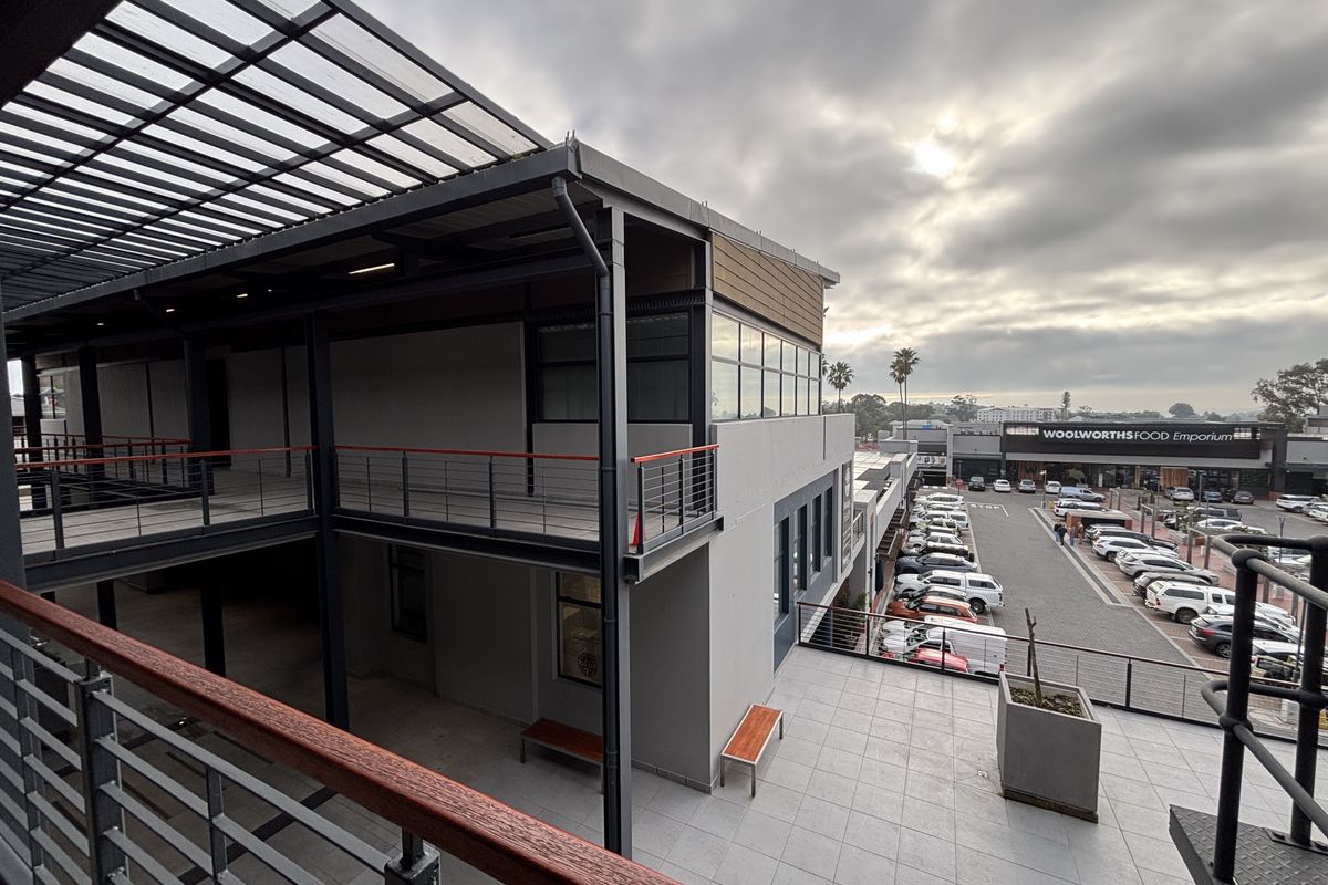 Modern multi-level office building exterior featuring a spacious tiled balcony, steel structural framing, and a view overlooking a commercial parking area.