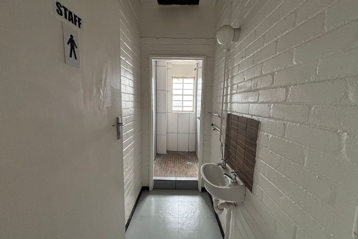A functional staff washroom area featuring a wall-mounted basin, painted brick walls, and a tiled shower cubicle beyond the doorway.
