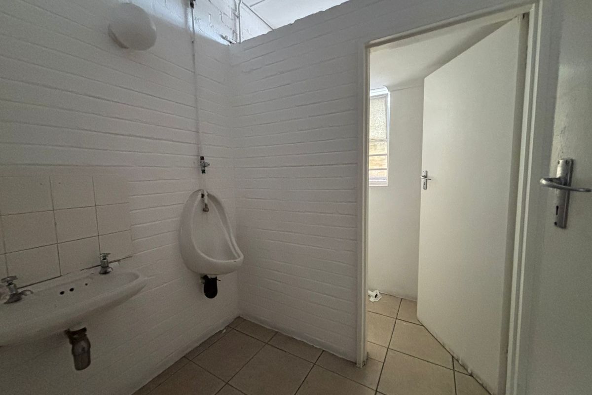 A functional industrial restroom facility featuring a wall-mounted urinal, a small basin, and tiled flooring with a separate partitioned area.
