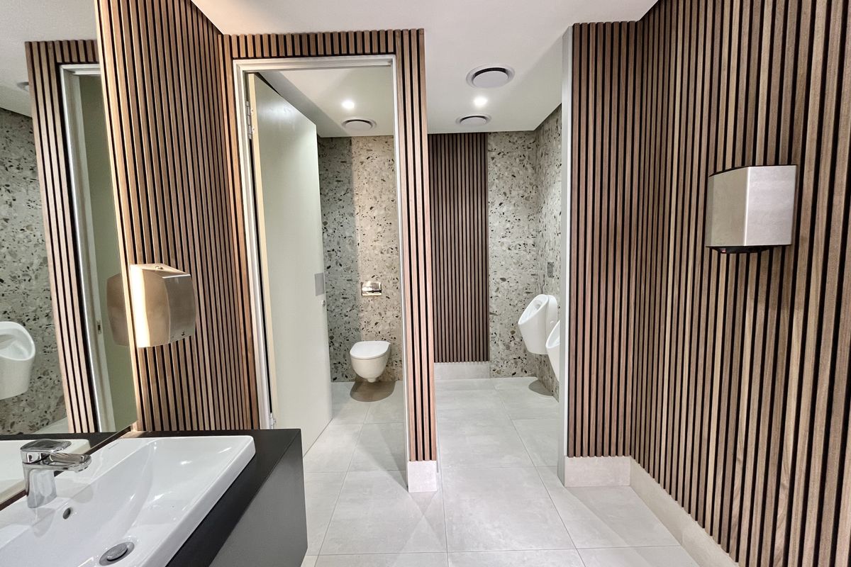 Modern office restroom featuring timber slat wall paneling, terrazzo-style wall tiles, contemporary sanitary ware, and recessed ceiling lighting.