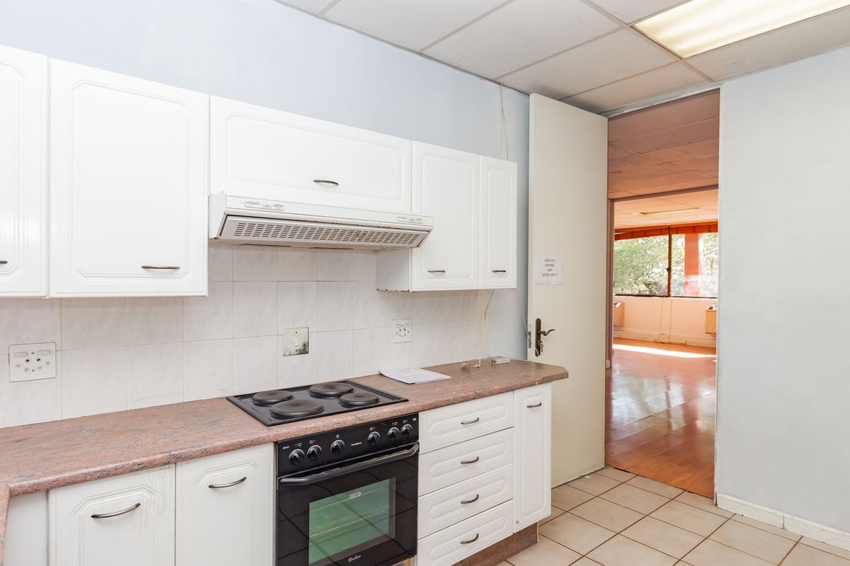 A functional office kitchenette featuring white cabinetry, a built-in oven and hob, and tiled flooring, with an adjacent open-plan office space visible.