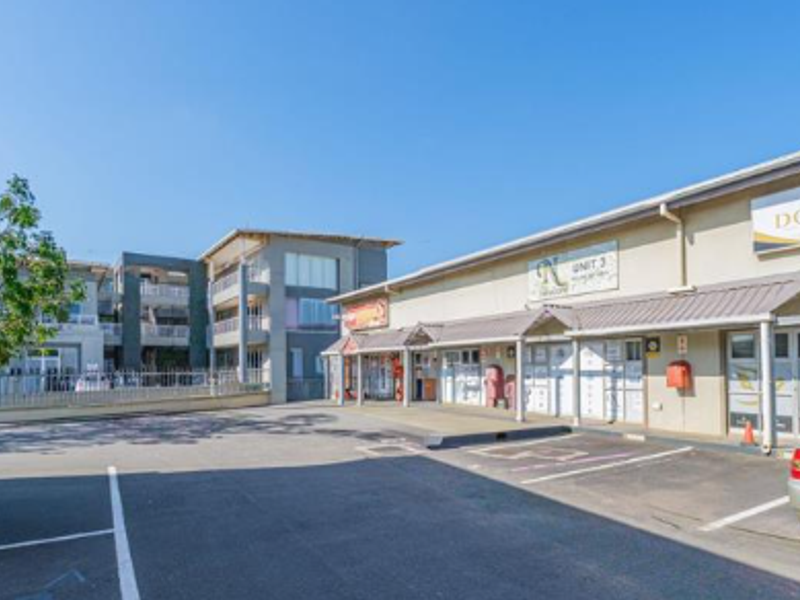Exterior view of a commercial complex featuring paved parking area, multiple unit entrances, and signage for various businesses.