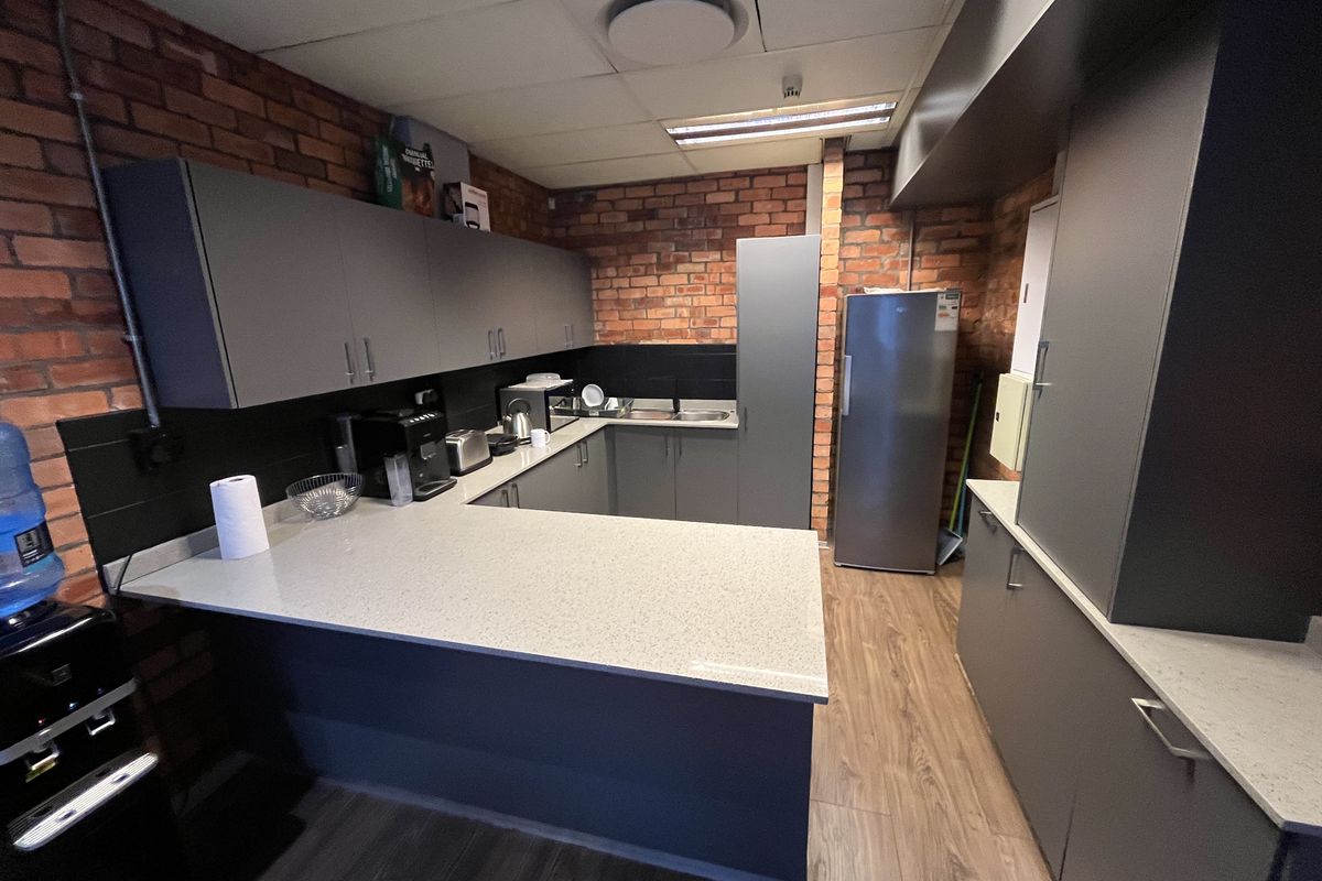 A modern office kitchenette featuring grey cabinetry, stone countertops, exposed brick walls, and a drop ceiling with integrated lighting.