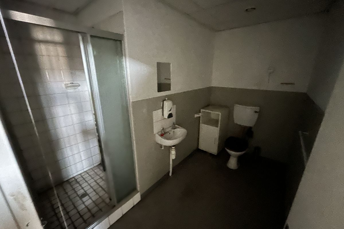 A functional industrial bathroom featuring a shower enclosure, wall-mounted basin, and toilet, requiring cosmetic updates to improve its overall condition.