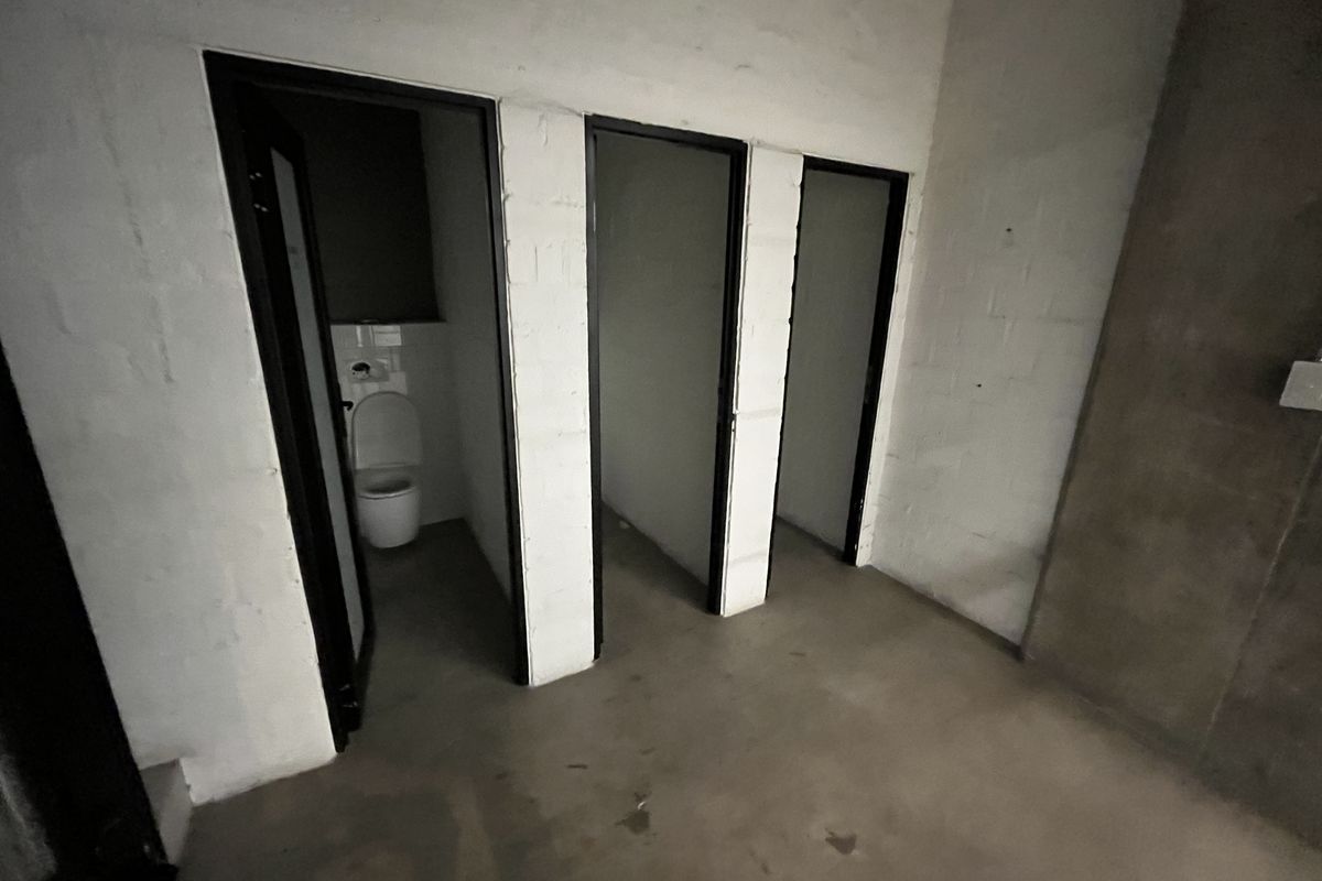A utilitarian restroom area featuring multiple toilet cubicles with exposed brick walls and concrete flooring, reflecting a basic industrial or unfinished aesthetic.