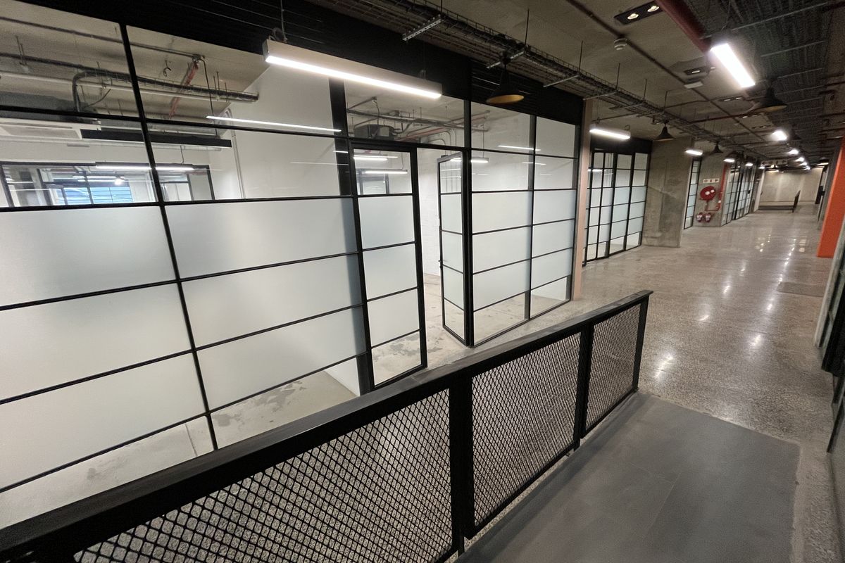 Modern industrial-style office corridor featuring polished concrete floors, exposed ceiling services, and black-framed glass partitioning for private office spaces.