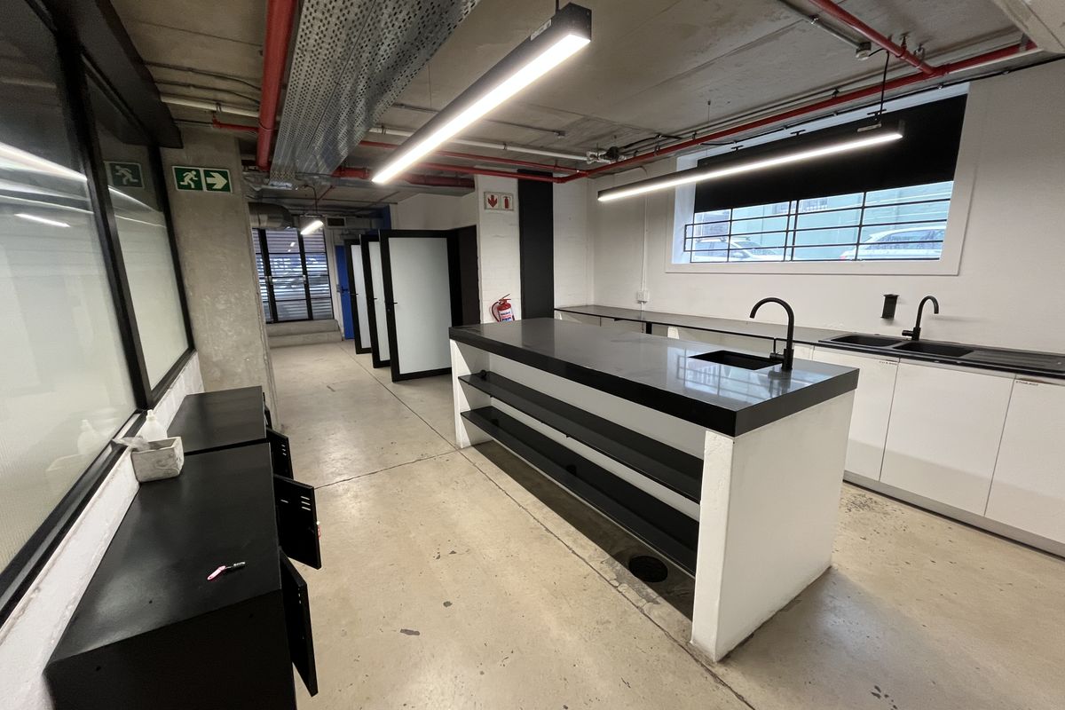 Modern industrial-style kitchenette featuring a central island with stone countertop, dual sinks, matte black fixtures, and exposed ceiling services.