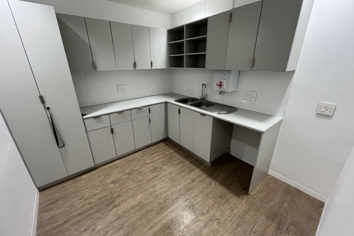 A modern office kitchenette featuring light grey cabinetry, laminate countertops, a stainless steel sink, and wood-look flooring.