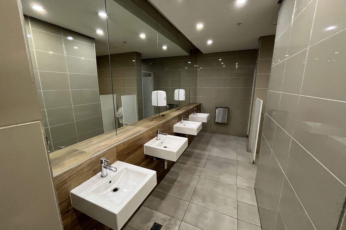 Modern commercial restroom featuring multiple wall-mounted ceramic basins, large vanity mirror, recessed ceiling lighting, and neutral floor-to-ceiling tiling.