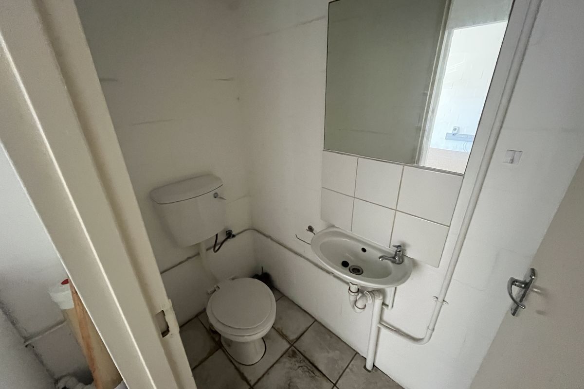 A compact industrial-style restroom featuring a standard toilet, wall-mounted basin, and mirror, requiring cosmetic updates to improve its overall condition.