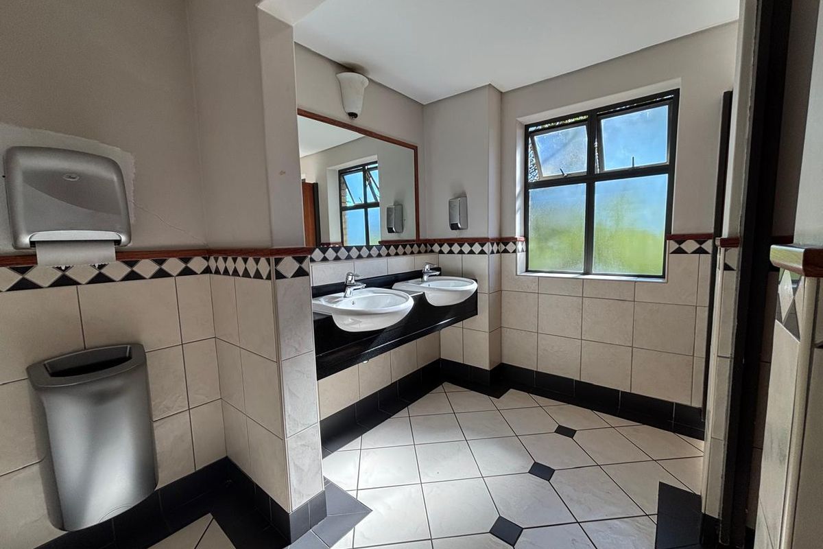 A functional office restroom featuring dual ceramic basins, a large vanity mirror, tiled walls with decorative border, and tiled flooring.