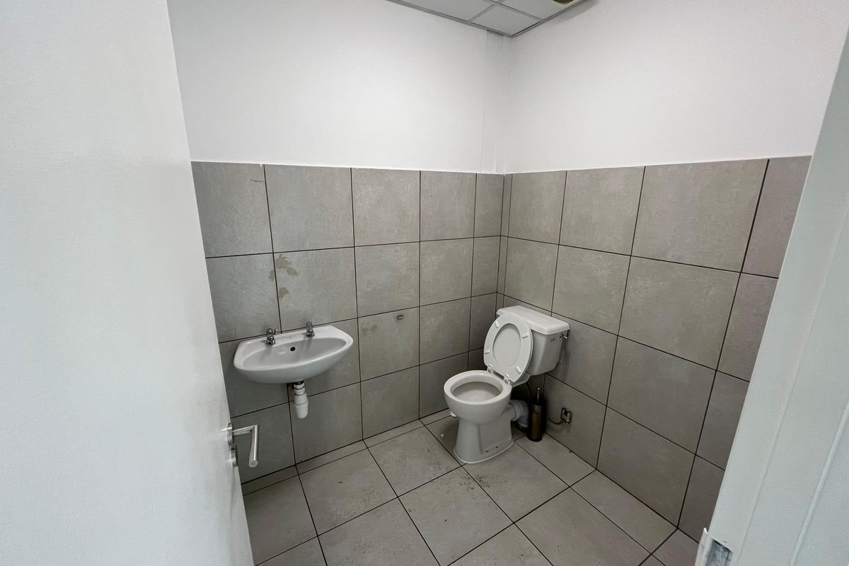 A functional industrial restroom featuring grey wall and floor tiling, a wall-mounted basin, and a standard toilet suite.